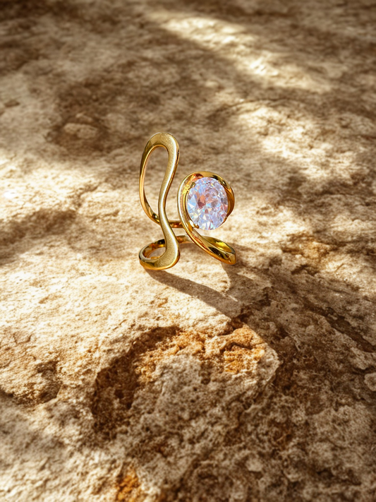 Sculptural gold ring with a large round cubic zirconia crystal, resting on sunlit textured stone. Mediterranean-inspired editorial setting with warm natural light and organic shadows.